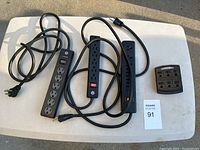 Photo of 3 different black power strips and one 6-outlet to 2-plug adapter on a white surface.