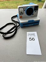 Front right angled view of the white and blue Polaroid One Step 2 instant camera with attached black wrist strap, sitting on a flat surface outside.