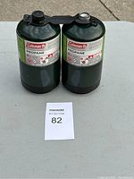 Two green Coleman propane canisters attached side by side on a light gray surface with a sale tag number 82 in front.