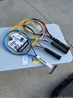 Four tennis racquets on white foldable plastic table: three Head racquets (blue/yellow 'Speed', yellow/black, silver/red) and one red Prince racquet, all with black grips.