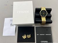 Citizen gold tone men's wristwatch displayed on black cushion stand with original white Citizen box, extra links, and instruction manual booklet included.