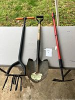 Full view of three yard tools laid out on a table showing a black pronged cultivator, a shovel with wooden handle, and a red-handled hoe tool.