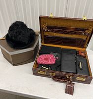 Open briefcase showing wallets and bags inside along with two vintage hats with their boxes and a wooden-handled parasol lying next to them.
