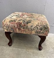 Angle of the foot stool showing floral fabric upholstery and curved wooden leg with polished finish.
