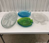 Top view of two large clear glass lobster plates, one large clear glass fish platter, five smaller fish-shaped glass plates in blue, green, and clear colors arranged on white table