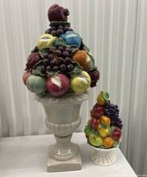 Full view of two ceramic fruit busts, large on left with urn base and small on right with pedestal base, showing multicolored ceramic grapes and other fruits