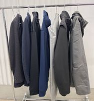View of seven men's jackets and one pair of pants on hangers on a rack showing a range of coats in black, navy, gray.