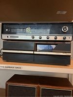 Front view of vintage 8-track player with multiple control knobs and two black speakers below it on shelving.