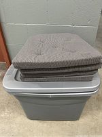 Four Ikea seat cushions stacked on top of a closed gray plastic storage tote.