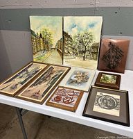 Overview of eight framed art pieces arranged on a table showing two watercolor paintings, multiple prints, and a wooden relief artwork.