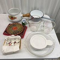 Overview showing all kitchenware items together on table including sauce pan, measuring cups, trivets, pie dish, and shot glasses