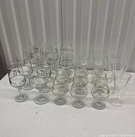 Grouped photo showing 24 clear glass stemware with gold decoration in rows on a white surface under soft light.