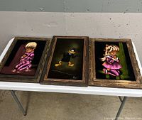 Three framed velvet paintings displayed on a white folding table, showing cartoon-style children praying on the left and right, with a Daffy Duck velvet painting in the center.