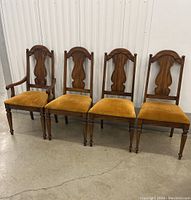 Set of four vintage wooden dining chairs, three without arms and one with armrests, dark wood with carved decorative backs and mustard upholstered seats, standing against a white wall on concrete floor.