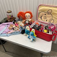 Overview photo showing various vintage dolls, toys, including a gumball machine, Raggedy Ann dolls, Smurf plushies, Barbie doll, and Winnie the Pooh suitcase with toys inside.