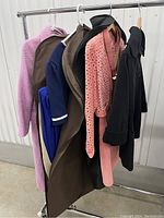 Photo of seven women's clothing items including a purple coat, tan suede set, coral pink sweater, dark navy and royal blue dresses, and a black garment hung on hangers on a clothing rack.
