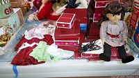 Overview of doll seated on table with red star boxes, wire hangers, dress pieces