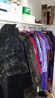 Hanging clothes showing black and brown fur coats along with other coats and dresses in various colors on hangers in closet.