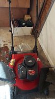 Top front view of the red Yard Machines lawn mower with handle and side discharge chute visible, placed indoors on a concrete floor.