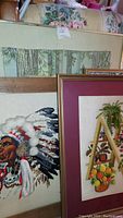 Several framed needlepoint artworks leaning against a couch with floral upholstery, showing varied designs including Native American chief and flowers.