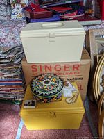 Looking at sewing boxes stack with decorative floral tin can and scattered small sewing items on top. Singer sewing machine box visible underneath.