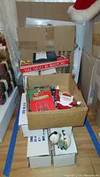 Photo showing stacked white boxes and open cardboard box containing various Christmas ornaments including Santa face and snowman figures.