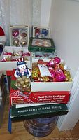 Photo showing multiple boxes of vintage glass Christmas ornaments in various shapes, colors, and packaging, along with boxed musical bells and a wooden Santa sleigh decoration.