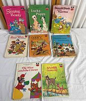Photo of eight Disney children's books laid out on white fabric. Titles include Sleeping Beauty, Lucky Puppy, Donald Duck and the Magic Stick, Snow White and the Seven Dwarfs, Goofy and the Milkman, Alice in Wonderland, The Mice and the Circus, and Bambi Grows Up. Books show signs of heavy use and some repaired damage.