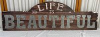 Full front view of the 'Life Is Beautiful' sign showing metal letters on textured cardboard backing with visible wear and damage.