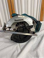Side view of Makita cordless circular saw showing the blade cover and handle with battery slot, teal and silver body with some wear and dirt.
