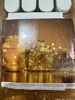 Close-up of packaging label showing warm white LED fairy lights wrapped around glass jars, with manufacturer information.