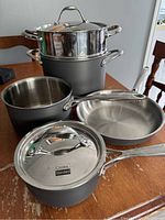 7 pieces of Cooks Standard cookware arranged on a wooden table including large 6L pot, steamer basket, saucepan with lid, frying pan and additional pots