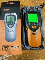 Digital stud finder device powered on, showing green screen display, placed next to its carry pouch and original retail box on wooden surface.