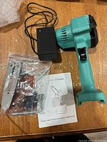 Handheld chainsaw body, power cord charger, plastic bag with blade, chain and hardware, and user manual on wooden surface.