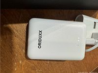 White Oriduxx branded powerbank with LED indicator lights and USB output ports, shown close-up on a wooden surface.