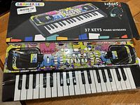 Front view of colorful Sanmersen 37-key electronic piano keyboard in original plastic wrapping with box.