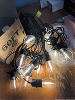 Photo of the string lights with clear elongated shatterproof LED bulbs on a black cable in front of packaging box.