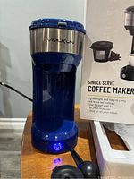 Front view of blue and stainless steel Vimukun single serve coffee maker with illuminated power button, shown next to its box.
