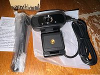Webcam shown mounted on tripod with USB cord coiled beside it, placed on a wooden surface with instructions sheet and part of the box visible.