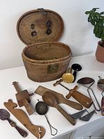 Open antique woven basket showing full lot of wooden hand tools and metal trinkets spread in front of the basket on a flat surface, basket positioned to show inside and front latch.