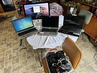 Full overview of the lot showing the iMac, three laptops, and a bin containing assorted cords and cables on a table with chairs around it.