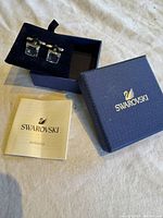Swarovski cuff links displayed on a black velvet cushion inside the original navy blue Swarovski box next to an instruction manual.