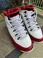 Pair of vintage Air Jordan sneakers placed outdoors on pavement showing front top and side view highlighting white and red color pattern, laces, and mesh tongue.