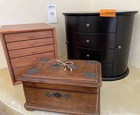 Three wood jewelry boxes arranged together: dark brown wood box with metal accents and key, black wood box with multiple drawers and side necklace cabinets, and light wood chest with drawers and inserts.