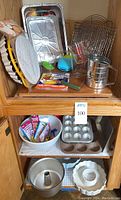 Shelving unit holding various metal baking pans such as upside down cake pan, bundt cake pan, muffin tin, cooling racks, measuring cups, and baking tools like sifters and spatulas.