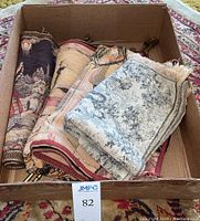 Four rolled and folded decorative tapestries in a cardboard box showing varying colors and designs.