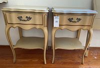 Pair of cream-colored French provincial style wood nightstands with curved legs, scalloped aprons and bottom shelves, each with one drawer with ornate metal pulls and gold accent lines.