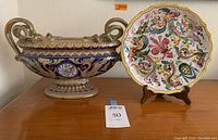 Photo of two Italian ceramics on wooden surface: an ornate oblong platter with handles and a round decorative plate on a stand
