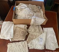 Box filled with folded lace and fabric table linens including various lace trims, napkins and doilies