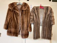 Two mink fur outerwear pieces hanging on hangers against a door, one brown and one natural grey mutation.